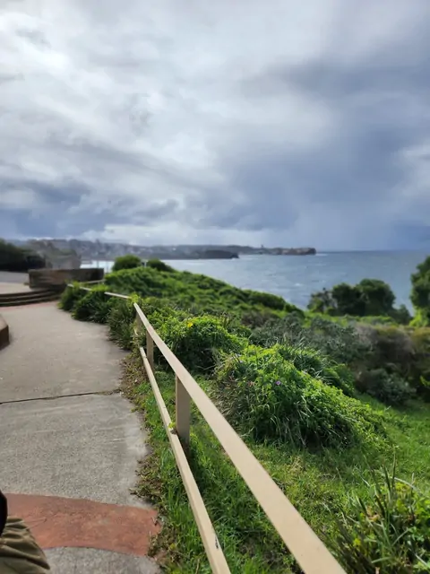 A coastal walking path leading toward the sea under a grey sky.