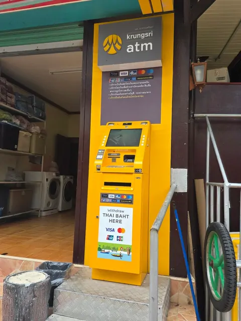 A yellow Krungsri Bank ATM located next to a shop's stairs.