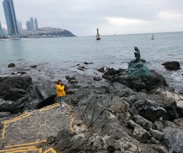 A traveler in a yellow jacket posing by the Mermaid statue at Haeundae.