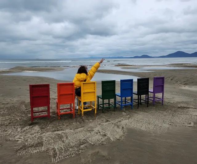 Rainbow-colored chairs on a grey beach with a woman in a yellow jumper.
