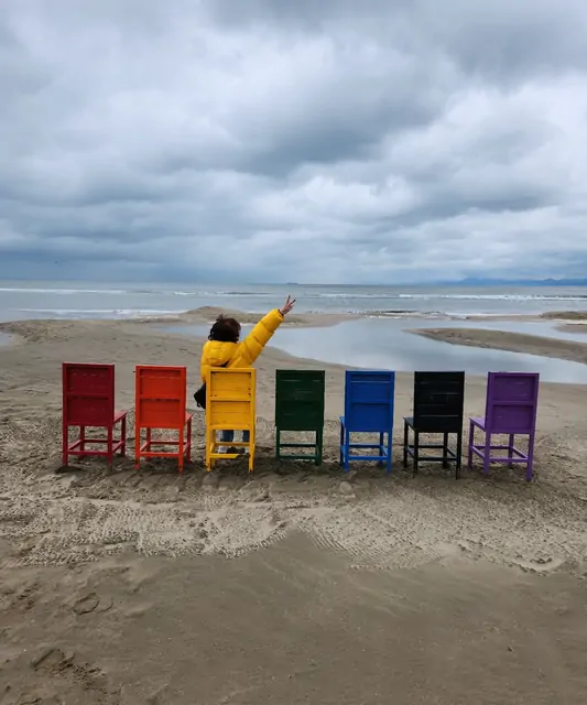 A row of colorful chairs on the sand as a woman poses for a photo.