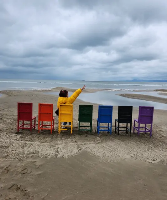 A woman in a yellow jacket pointing to the sky beside the rainbow chairs.