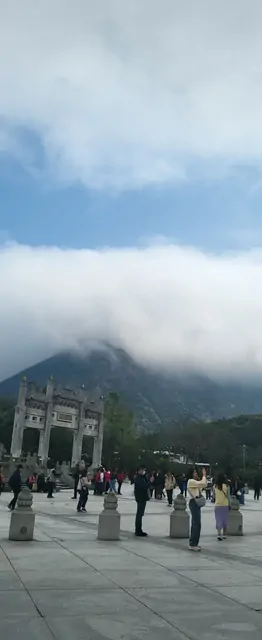 Tourists walking past a stone gateway with cloudy mountains in the background.